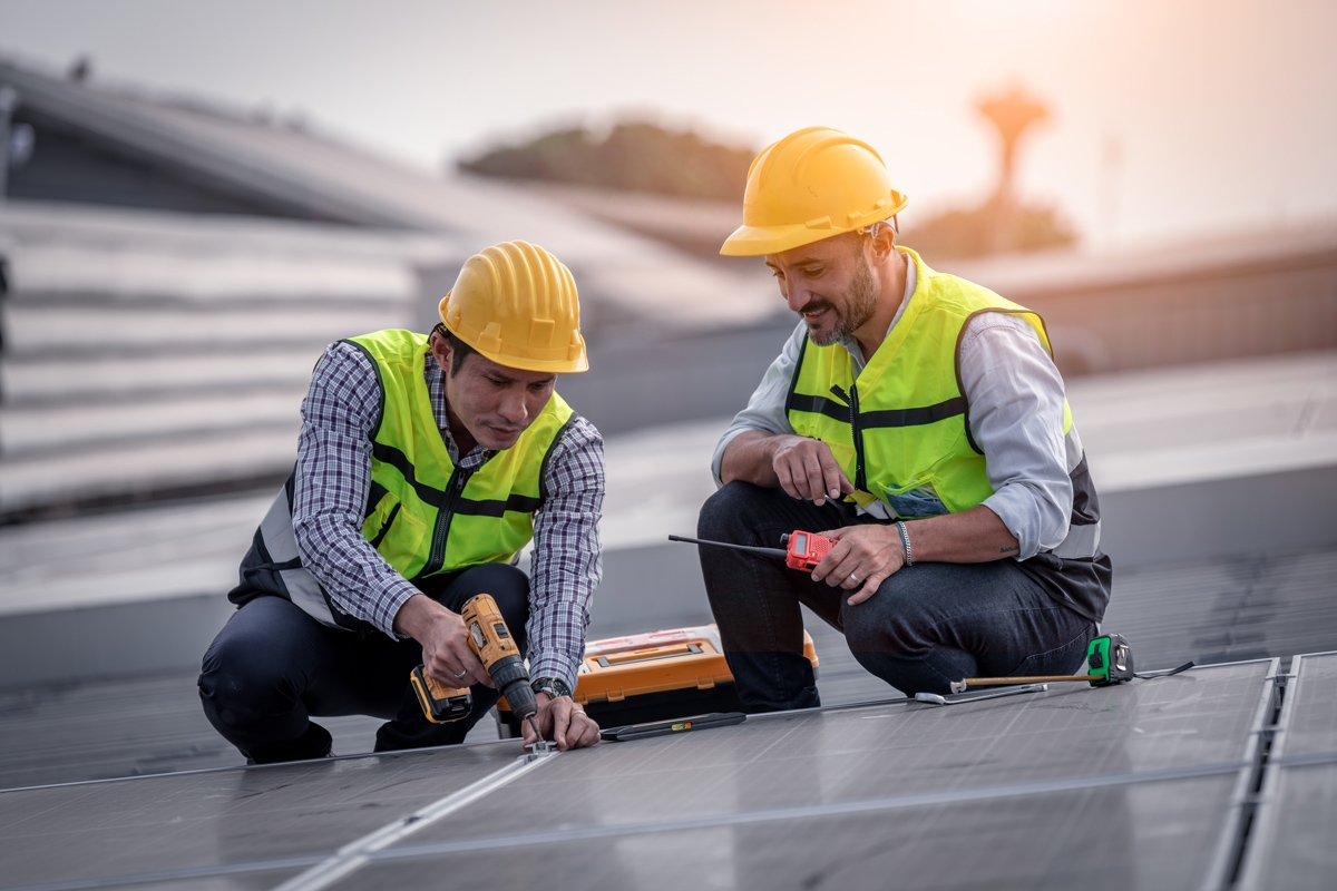 Two workers wearing safety helmets and high-visibility vests install solar panels on a rooftop using tools.