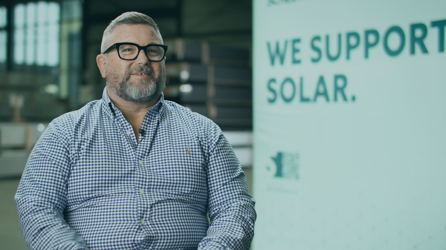 A man wearing glasses and a checkered shirt is seated in front of a sign that reads "We support solar.