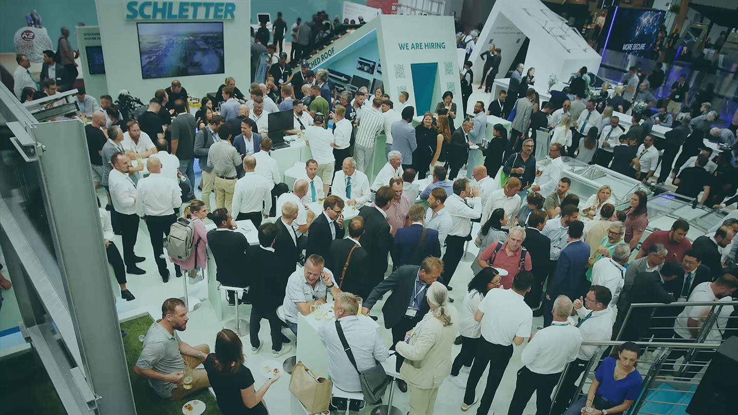 A large group of people attending a bustling trade show or expo, with various booths and displays in the background, and a banner that reads "Schletter" visible.