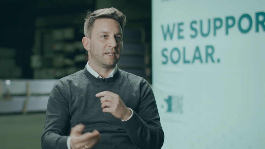 A man in a grey sweater and white shirt speaks while gesturing with his hands. A sign in the background reads "We support solar".