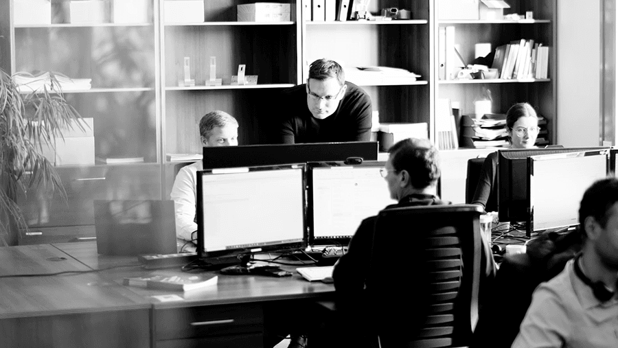 A black and white photo of four people working in an office. Two individuals are seated at desks with computer screens in front of them, while two others are standing and engaged in discussion.