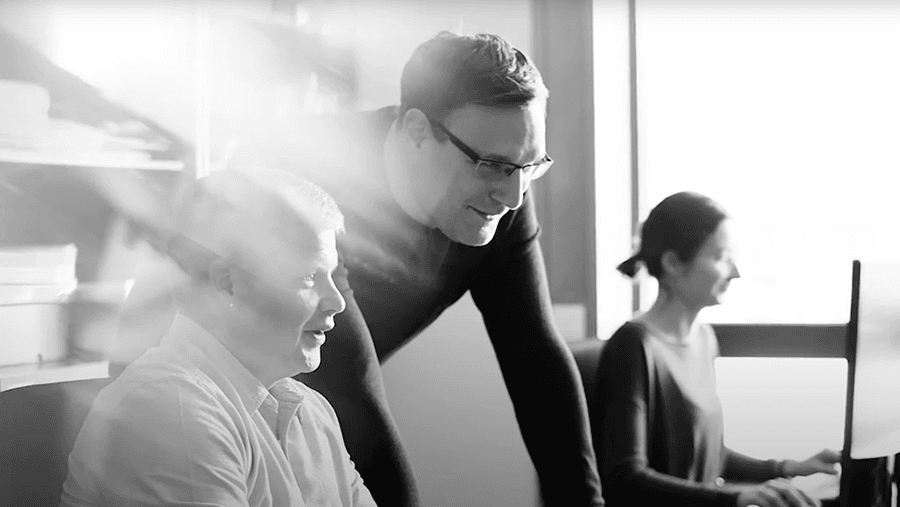 Three people are working in an office. One person stands, leaning forward, while two others sit at desks with computers. The atmosphere appears collaborative and engaged.