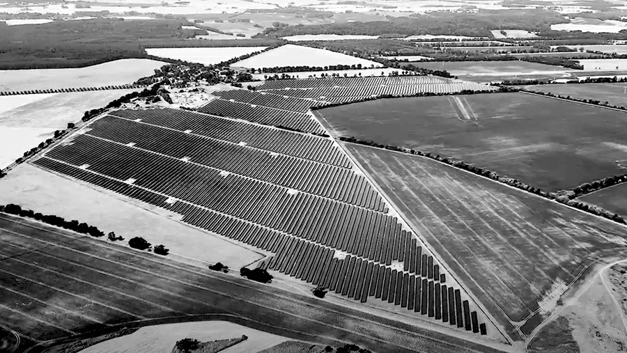 Aerial view of a large solar farm with rows of solar panels arranged in a triangular pattern surrounded by agricultural fields.