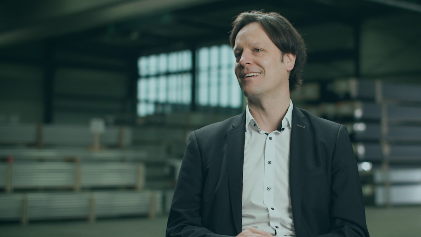 A man in a dark blazer and light shirt smiles while seated in a large industrial space with metal shelves and windows in the background.