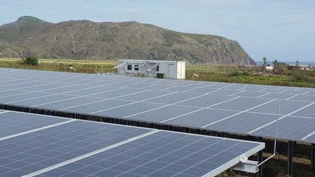 Large solar panel installation in a field with a small building in the background and a mountain range visible beyond.