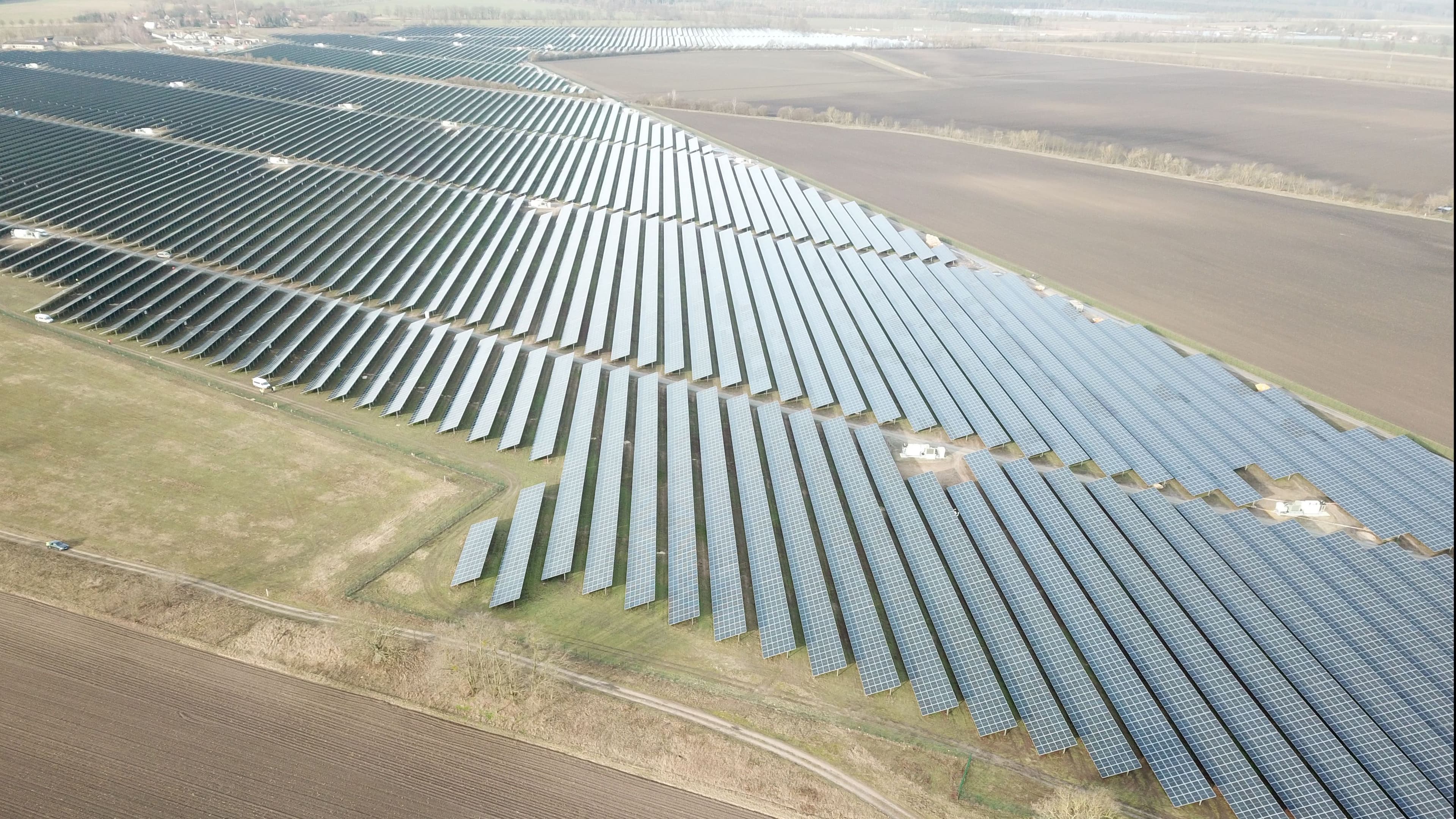 Aerial view of a large solar farm with rows of solar panels in a rural landscape. Fields and distant buildings are visible.
