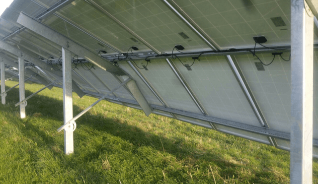 Underside view of solar panels mounted on metal frames in a grassy field. The image shows the wiring and structural supports.