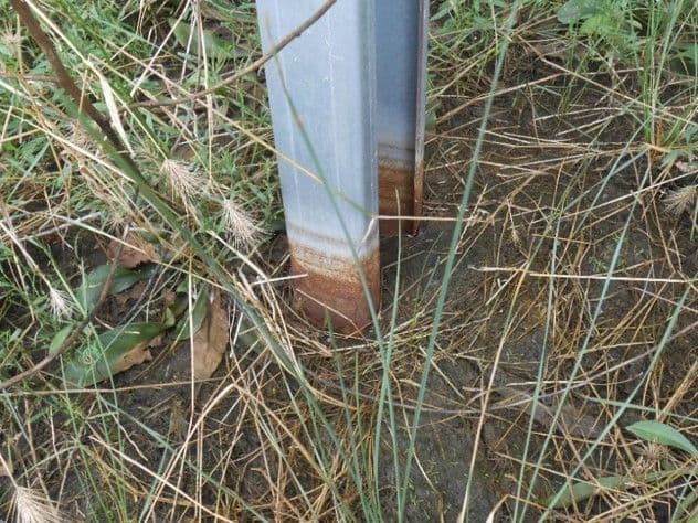 Close-up of a metal pole partially buried in soil and surrounded by dry grass, with visible rust at the base.