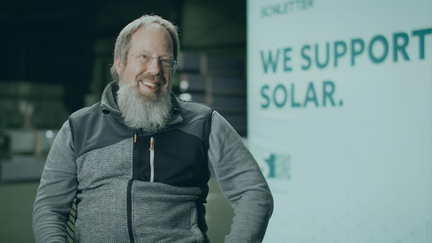 A man with glasses and a beard sits smiling in front of a sign that reads "WE SUPPORT SOLAR.