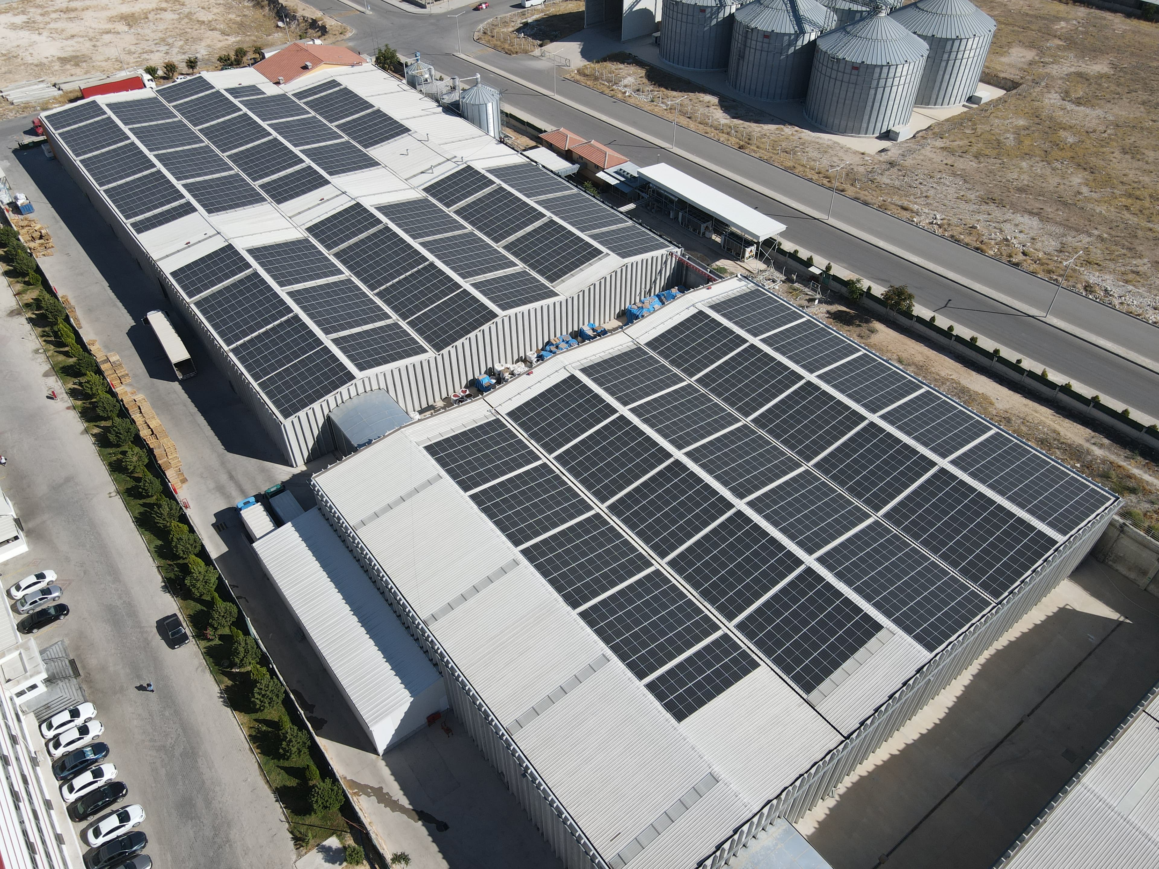 Aerial view of industrial buildings with large solar panels on the roofs, surrounded by paved areas and roads, with silos visible in the background.