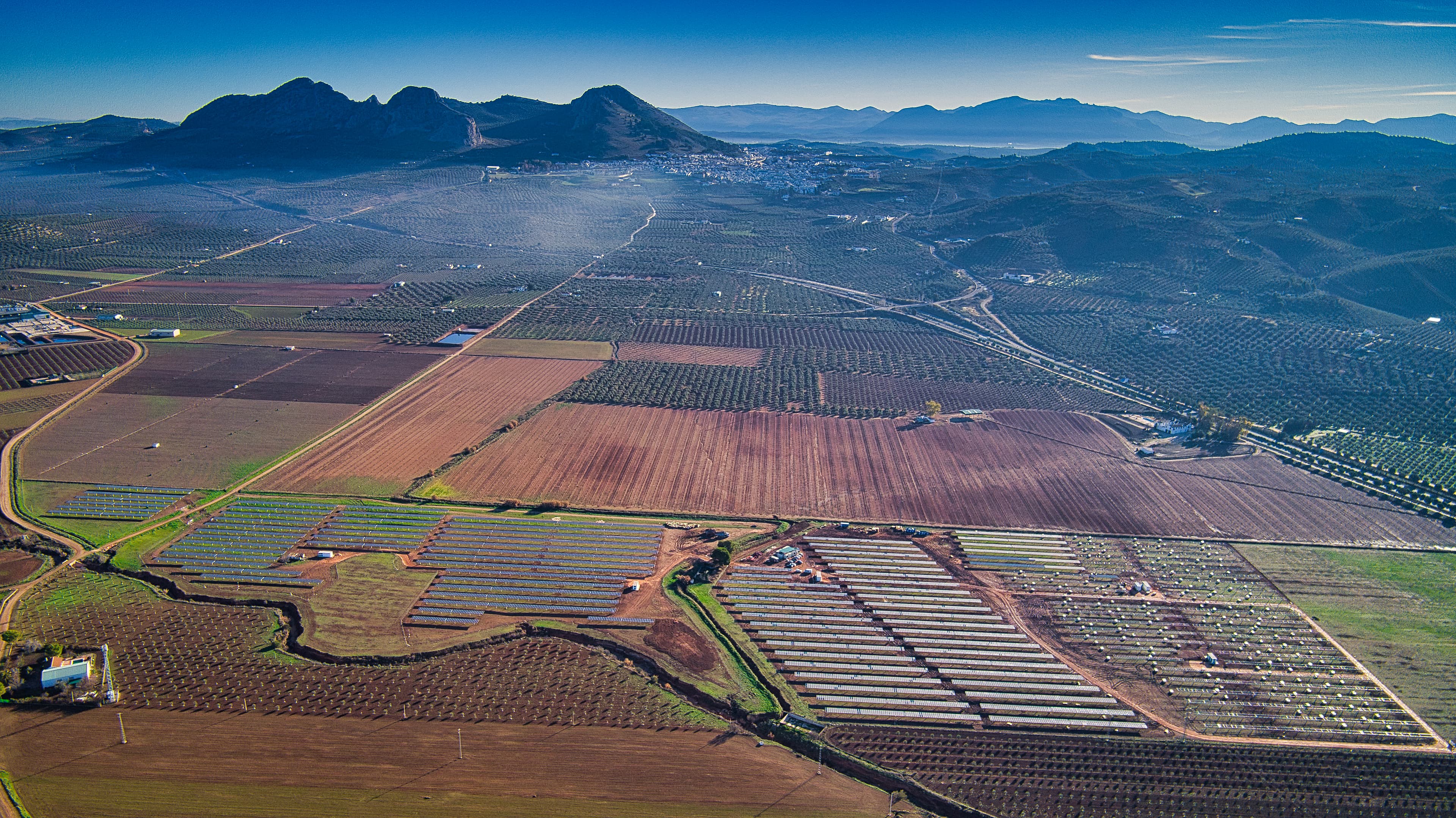 Aerial view of a landscape featuring agricultural fields, rows of crops, and mountains in the background under a clear blue sky.