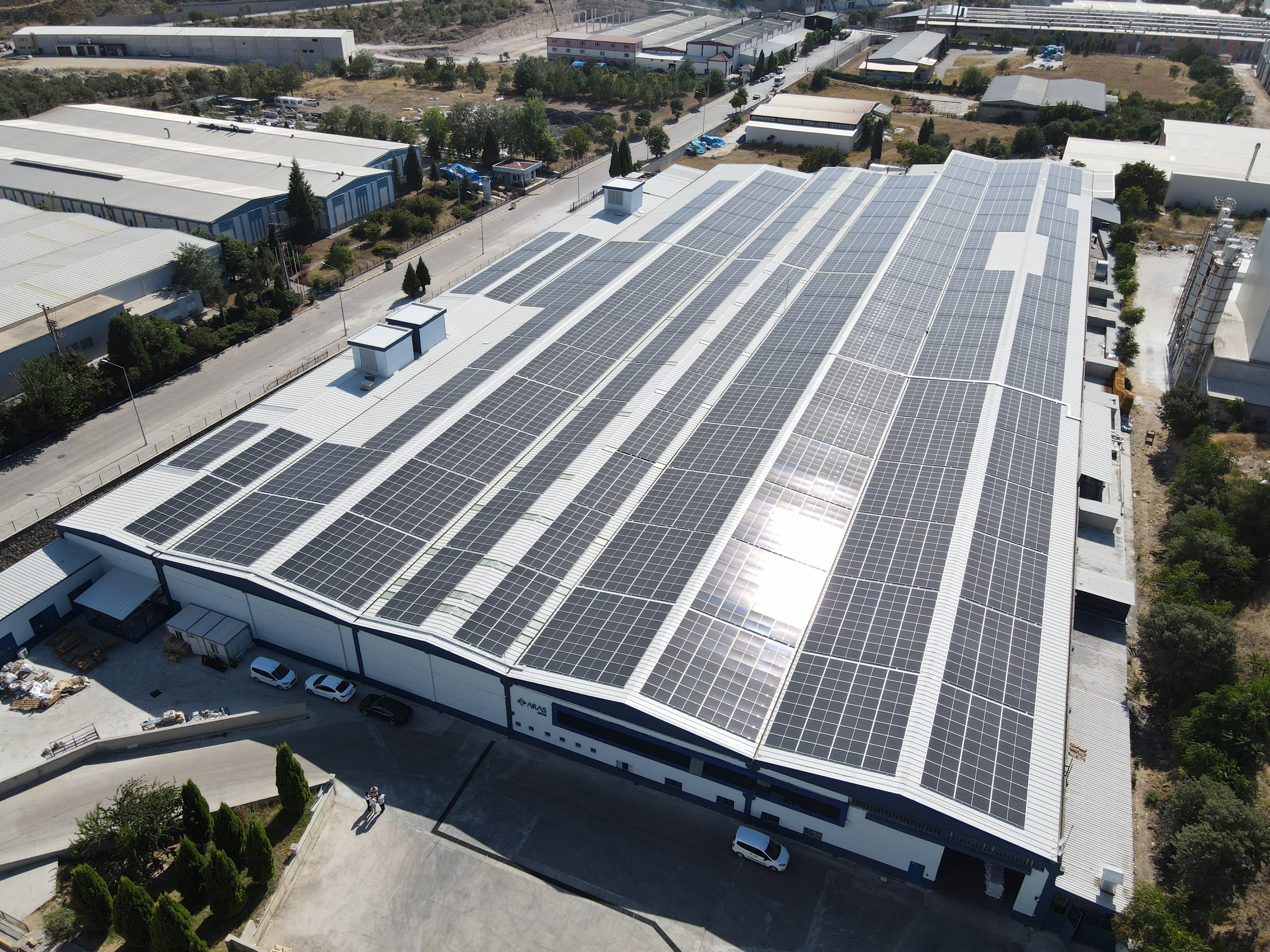 An aerial view of a large industrial building with solar panels covering its roof, situated in a commercial area with trees and other buildings nearby.