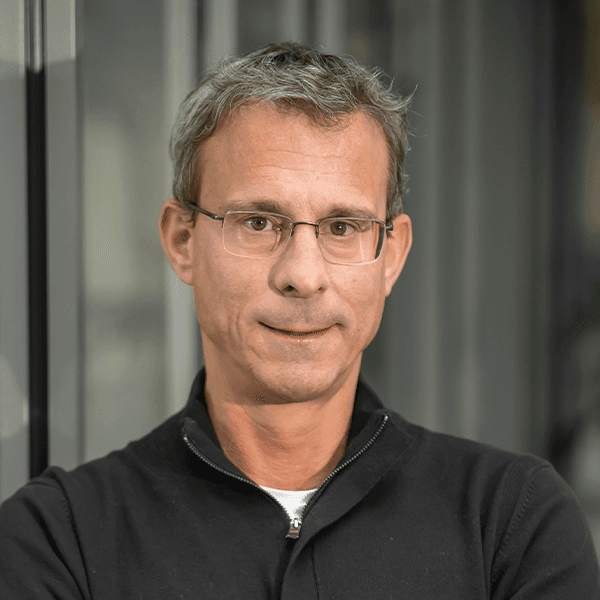 Man with short gray hair and glasses wearing a black zip-up jacket, standing against a blurred background.