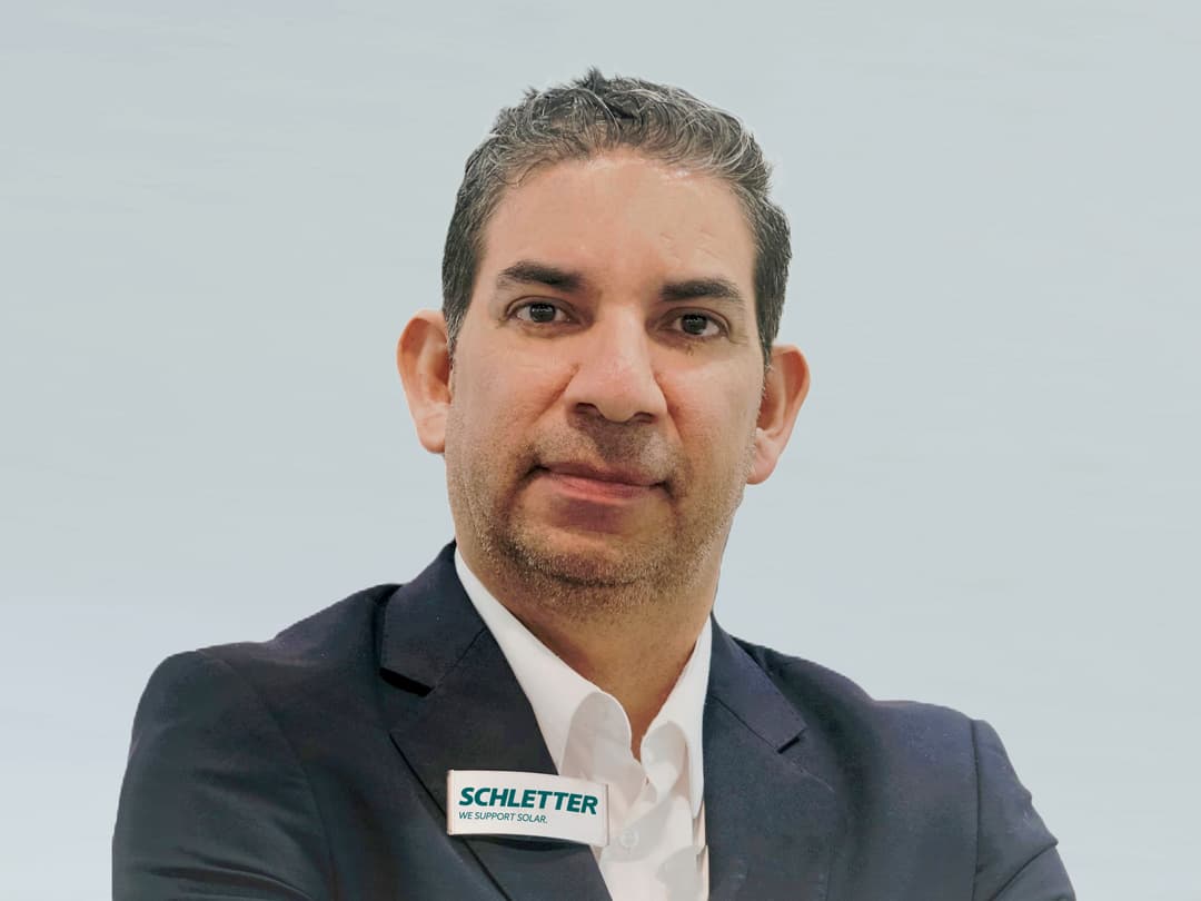 A man wearing a dark suit jacket and white shirt. He has short hair and a neutral expression, and a pin on his lapel with the text "SCHLETTER WE SUPPORT SOLAR.