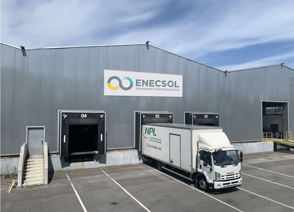 A large industrial building with a sign "ENESCOL Equipement Photovoltaïque." A white truck labeled "NPL" is parked at one of the loading docks. The building has four dock doors and an exterior staircase.