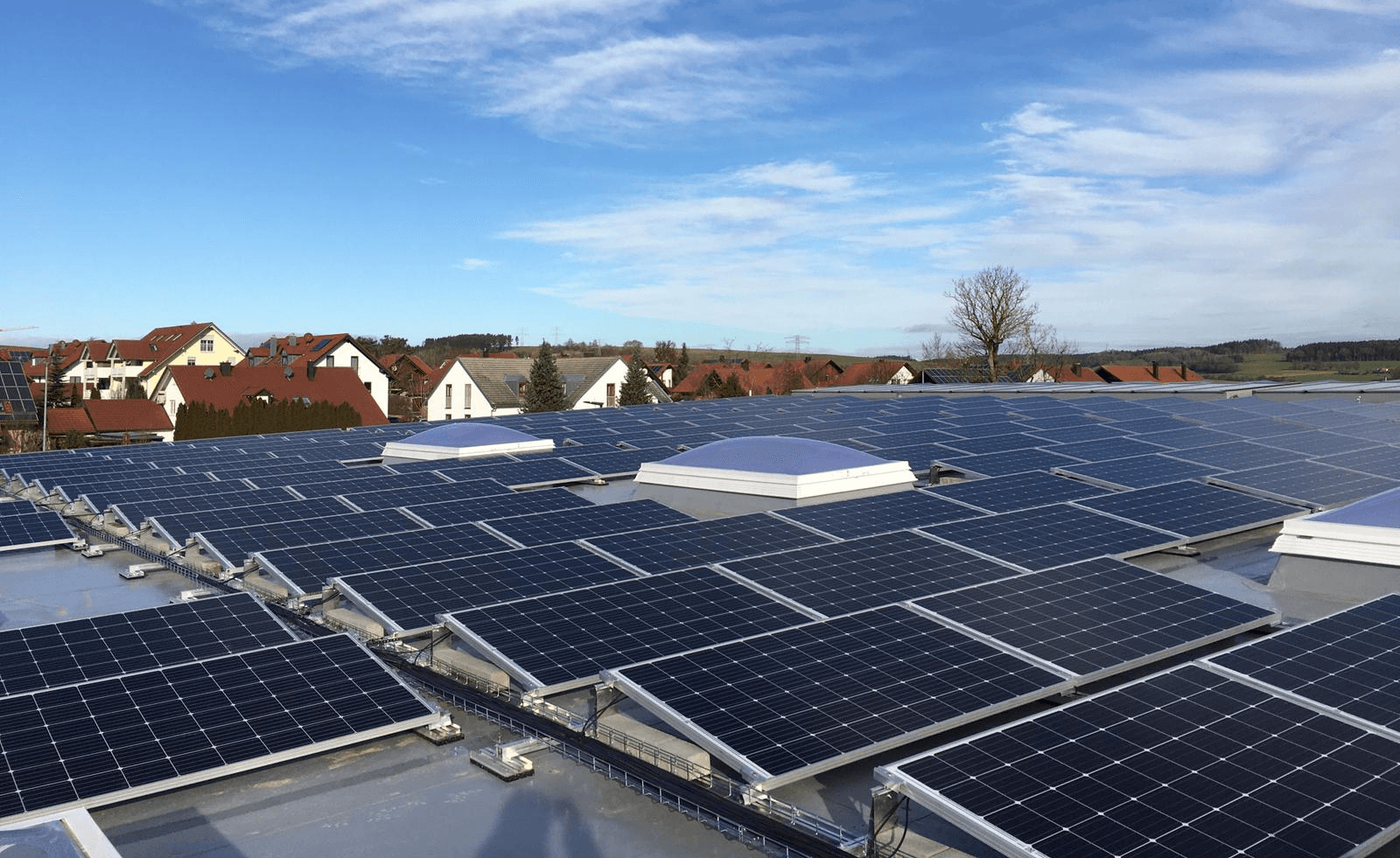 A rooftop covered with solar panels in a residential area with houses and a partially cloudy sky in the background.