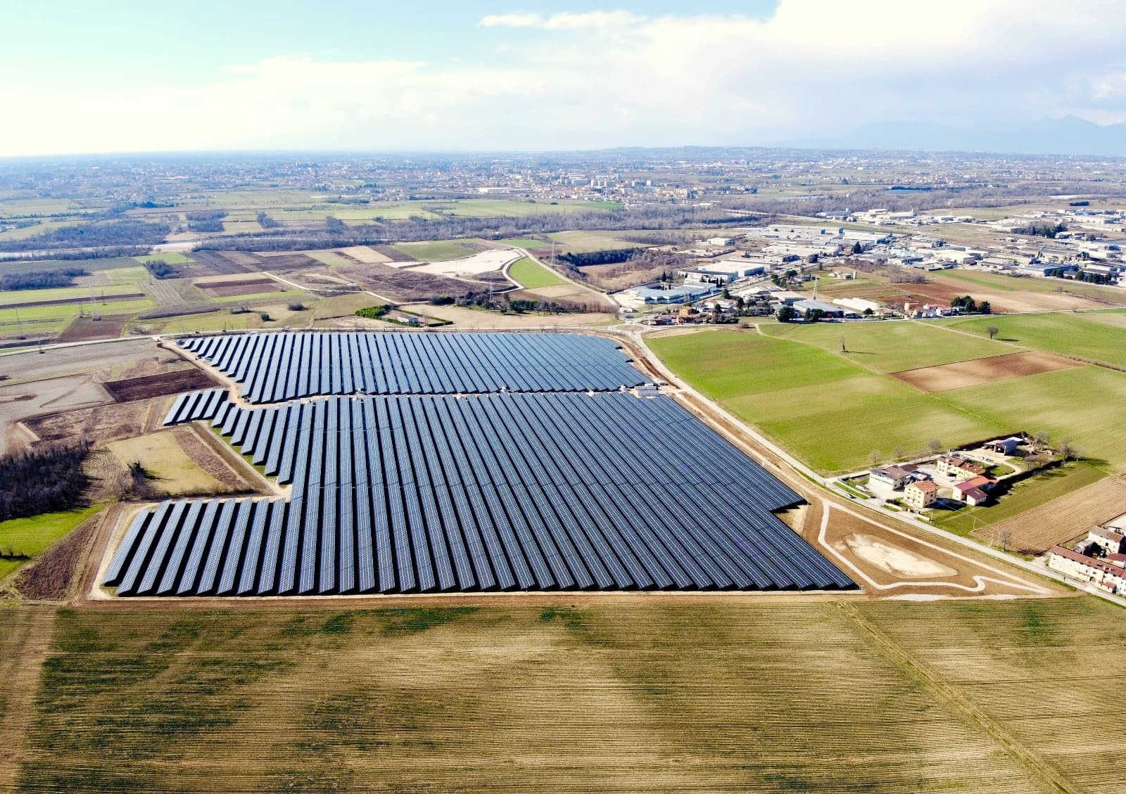 Aerial view of a large solar farm with multiple rows of photovoltaic panels situated in a rural area with fields, roads, and buildings visible in the background.