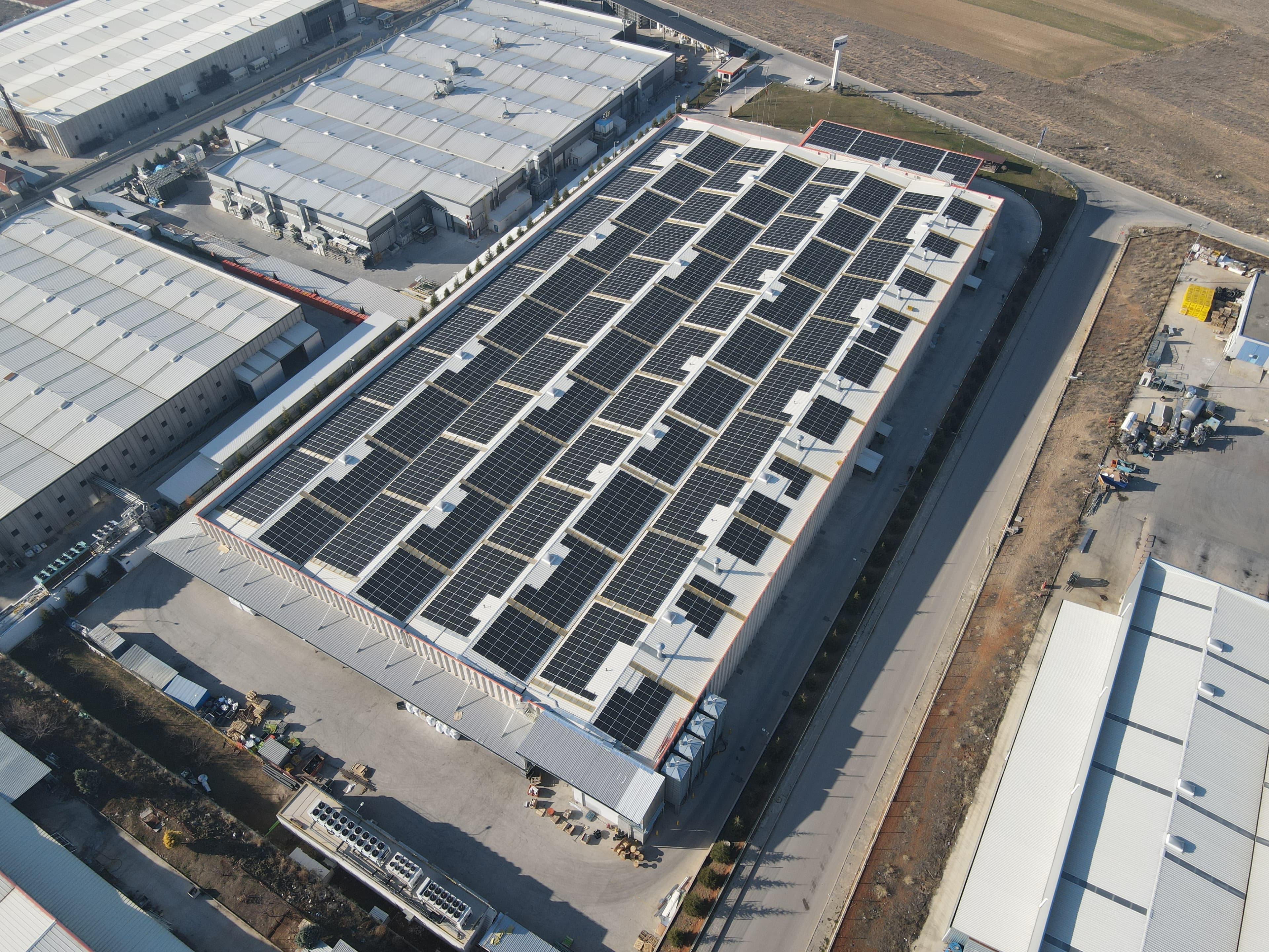 Aerial view of an industrial building with a large array of solar panels on its roof, surrounded by other industrial structures and a landscape with fields in the background.