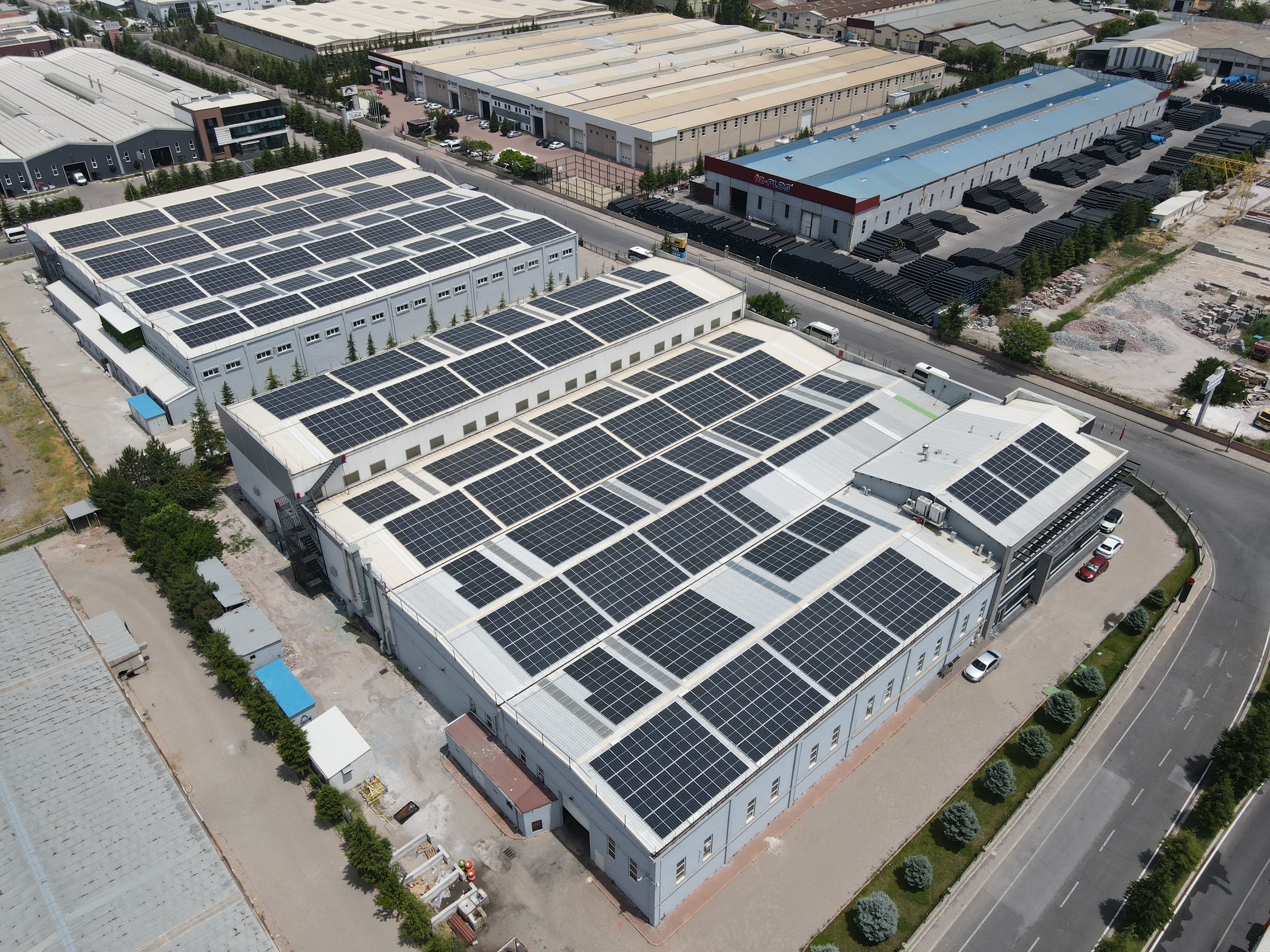 Aerial view of a large industrial complex with multiple buildings, all equipped with extensive solar panel installations on their roofs. Surrounding area includes roads and other industrial structures.