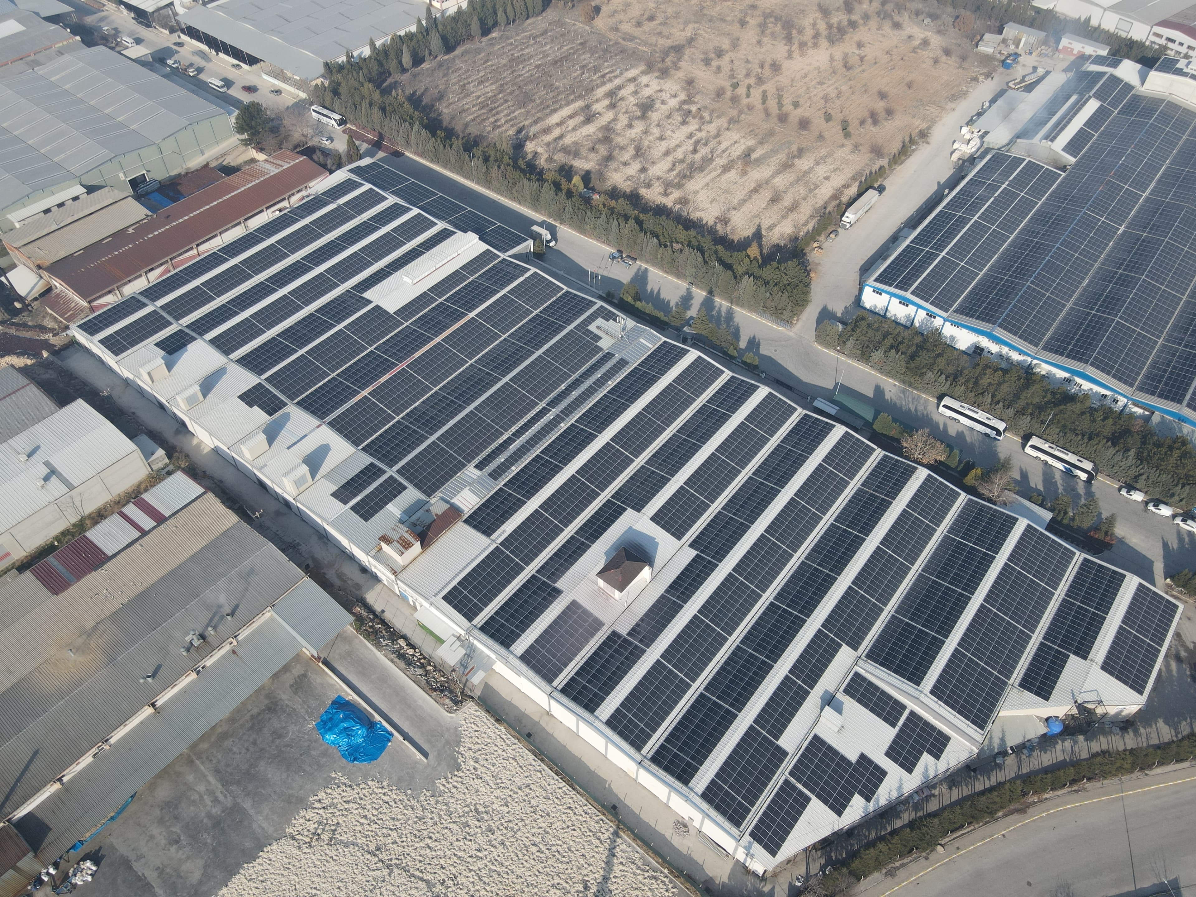 Aerial view of large industrial buildings with extensive rooftop solar panels, adjacent open land areas, and nearby parked buses.