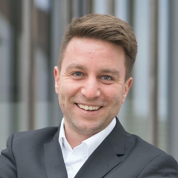 A man in a suit and white shirt smiles at the camera with crossed arms. The background shows vertical lines on a blurred building.