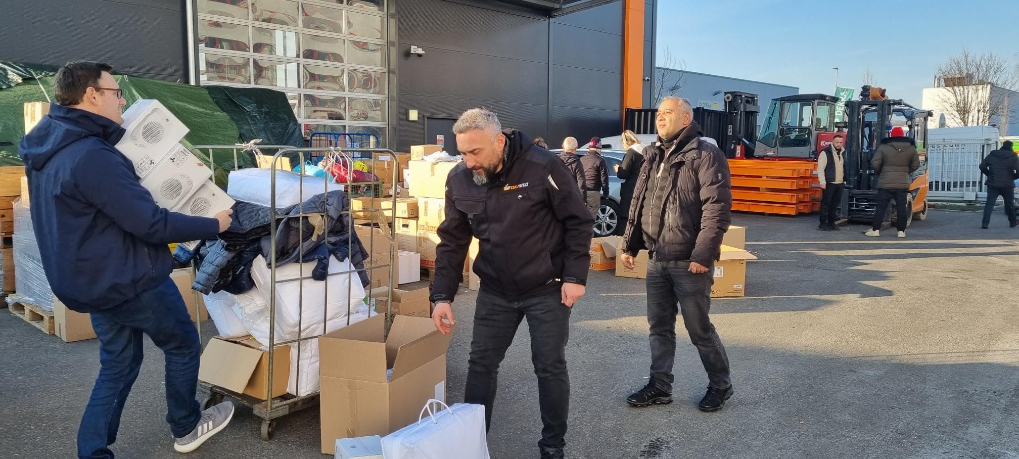 People unpacking boxes and equipment outdoors near a warehouse with forklifts and vehicles in the background.