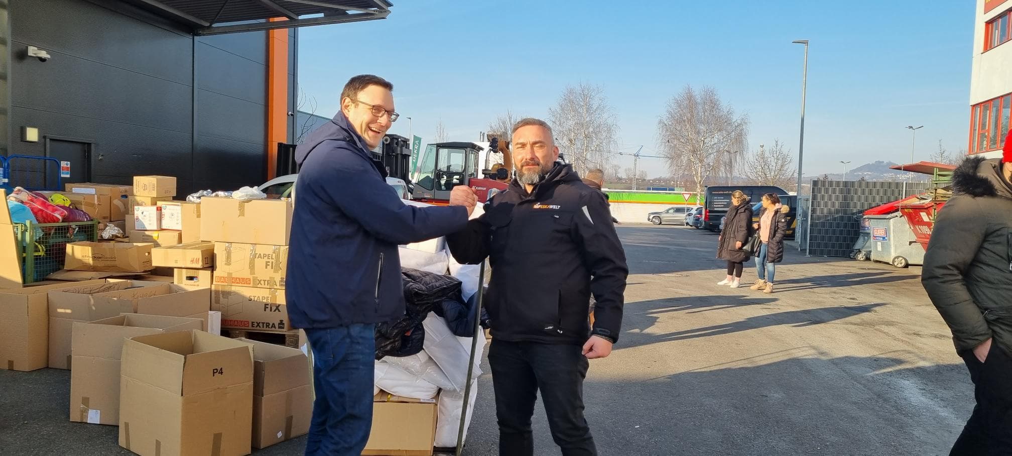 Two men shaking hands outside a building, surrounded by piles of boxes and bags. Other people are visible in the background near more boxes. The sky is clear and a forklift is parked nearby.