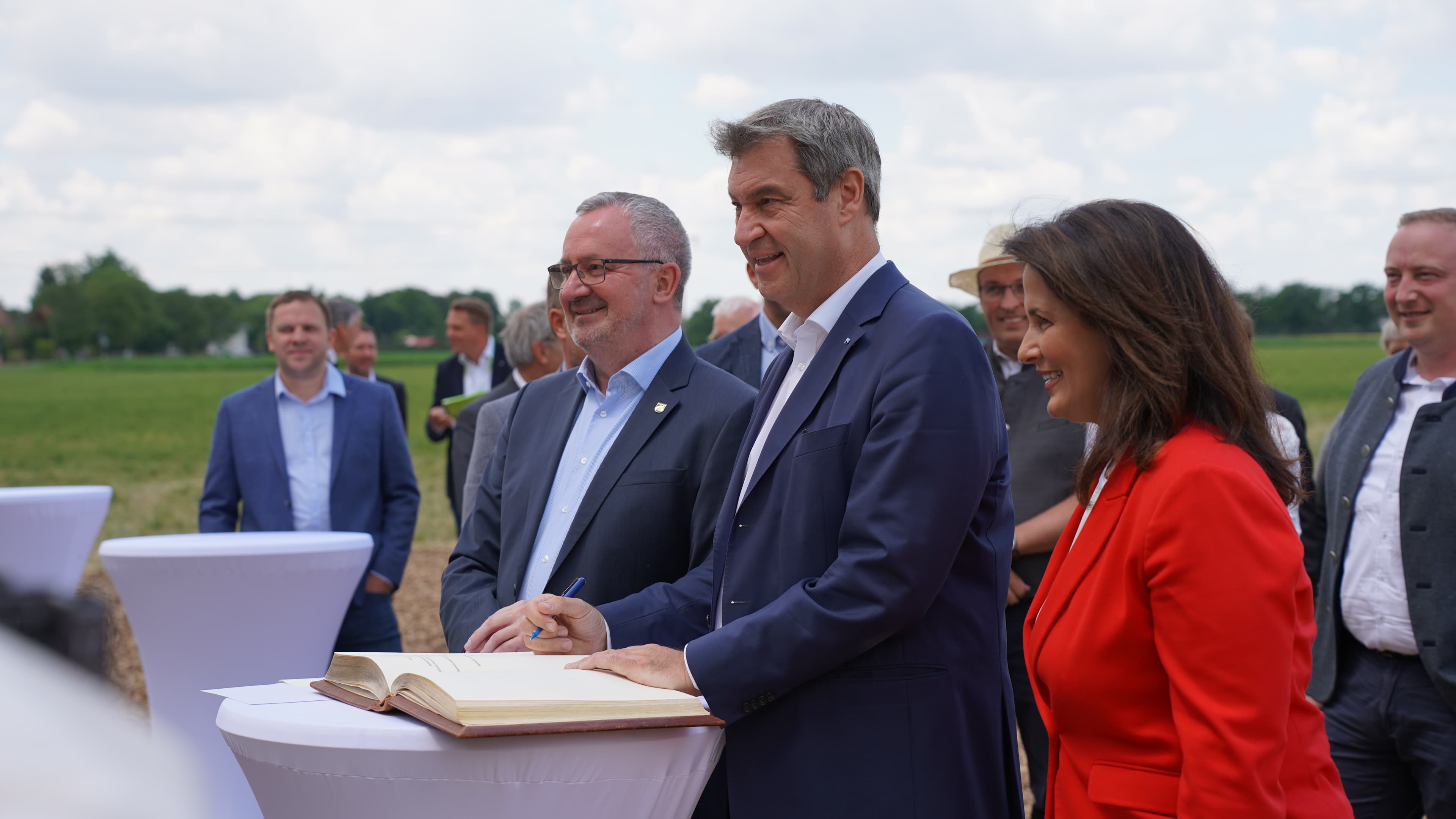 A group of people in business attire stand outdoors around a tall table, with one person in the center signing a document in a large book.