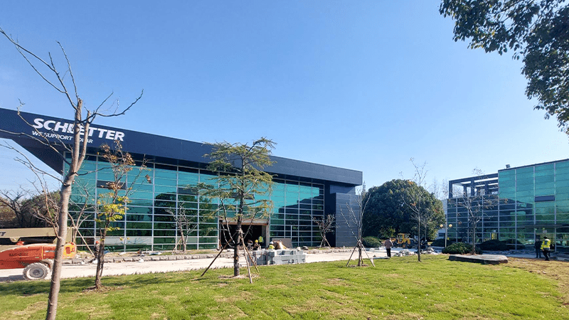 Modern glass office building with "SCHIETTER" signage on facade, surrounded by a well-manicured lawn and trees. Construction workers and equipment are seen in front of the building.