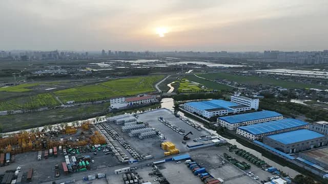 Aerial view of an industrial park with warehouses, containers, and equipment, surrounded by fields and a river under an overcast sky with the sun setting on the horizon.