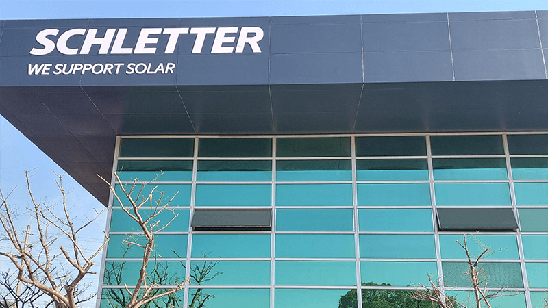 A building with the sign "Schletter We Support Solar" prominently displayed above large windows. Tree branches are visible in the foreground.