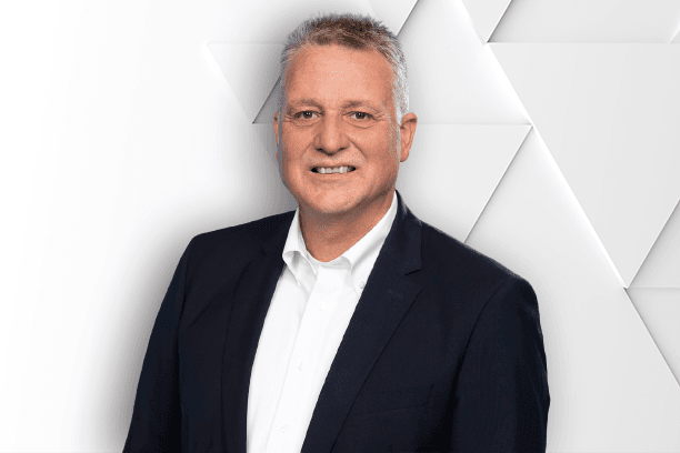 A man with short gray hair is wearing a dark suit jacket and a white shirt, standing against a geometric white background.