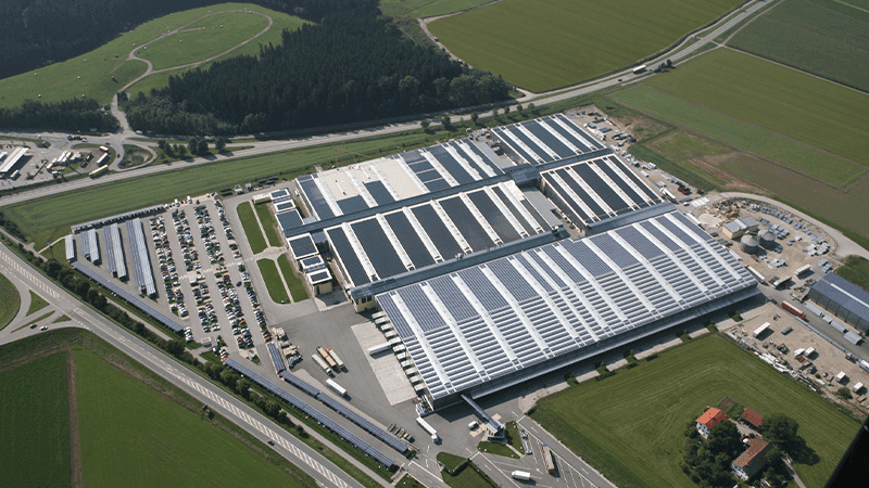 Aerial view of a large industrial complex with multiple buildings, surrounded by greenery and fields, featuring parking lots and access roads.