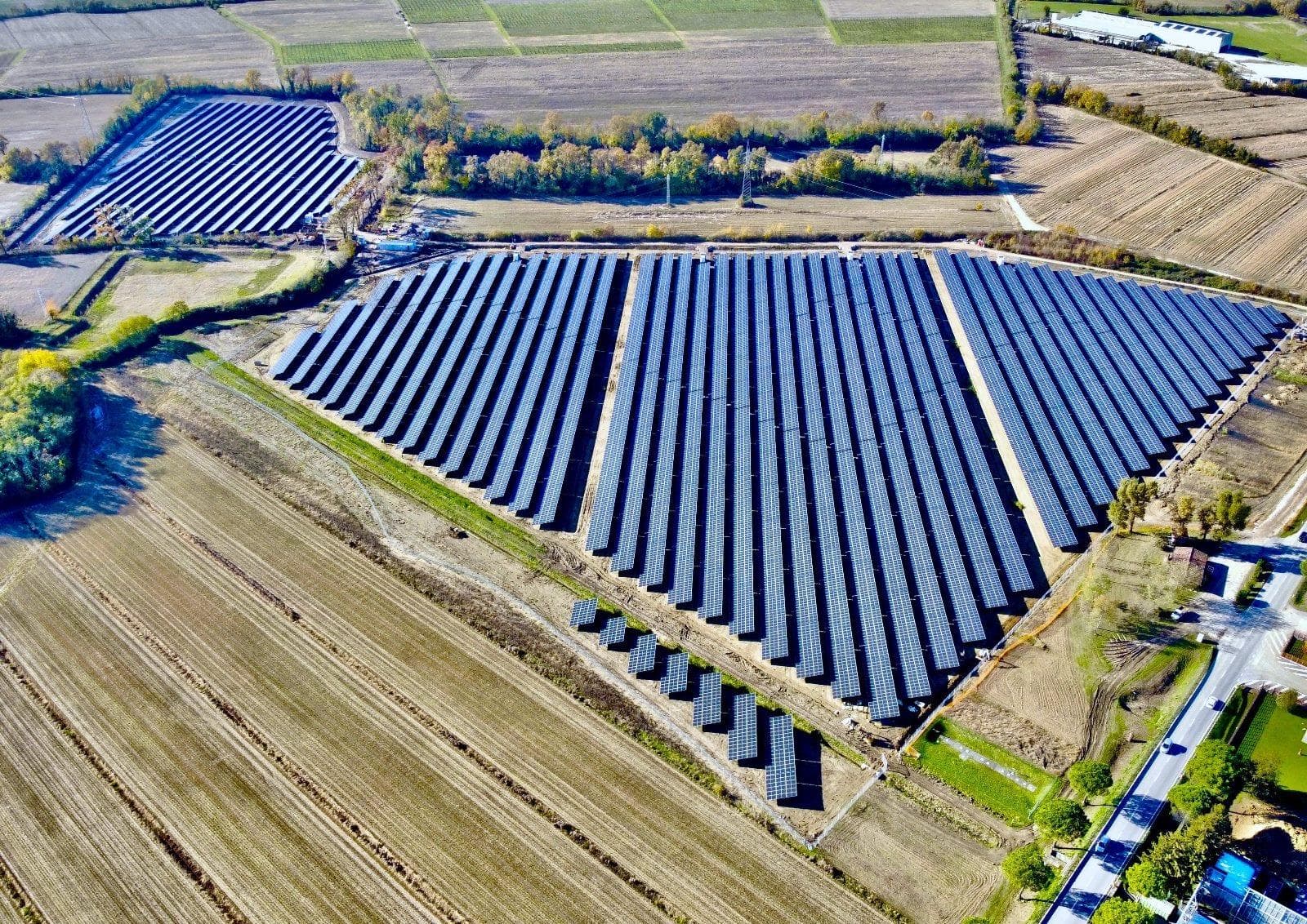 Aerial view of an expansive solar farm in a rural area, with large arrays of solar panels arranged in parallel rows amidst fields. Trees and additional farmland surround the installation.