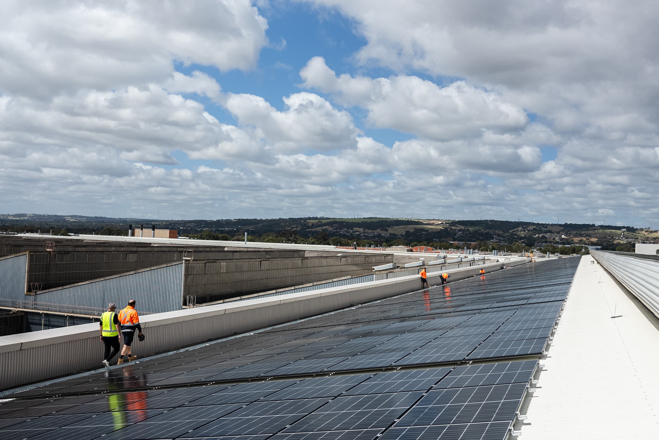 Workers in high-visibility vests walking on a large rooftop covered with solar panels under a partly cloudy sky.