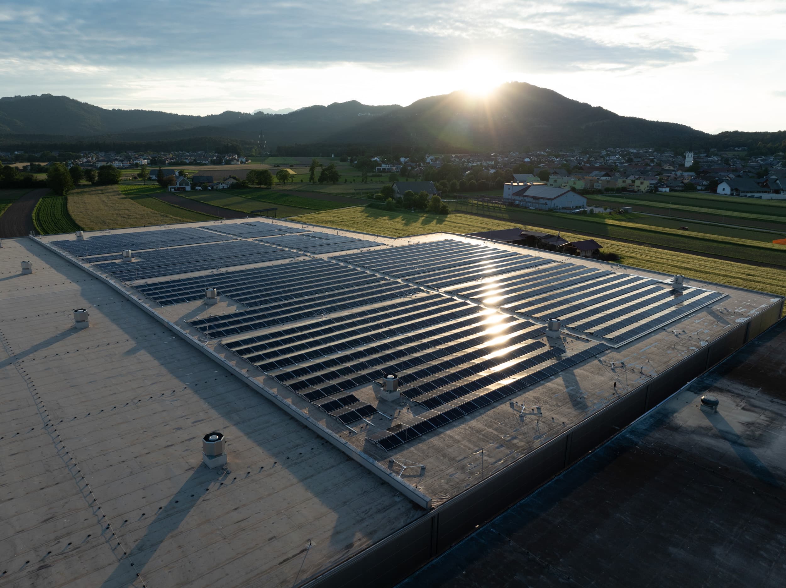 Sun rising over a facility with a large array of solar panels on its roof, set against a backdrop of fields and distant mountains.