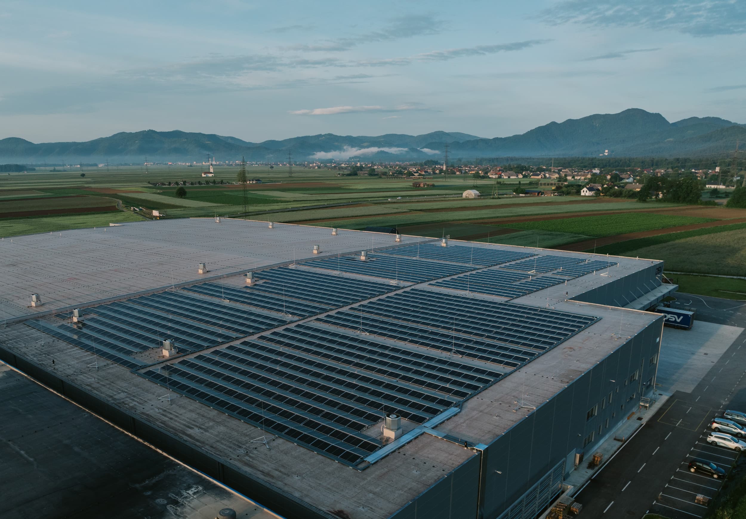Aerial view of a large industrial facility with solar panels on the roof, set in a rural area with fields and mountains in the background.