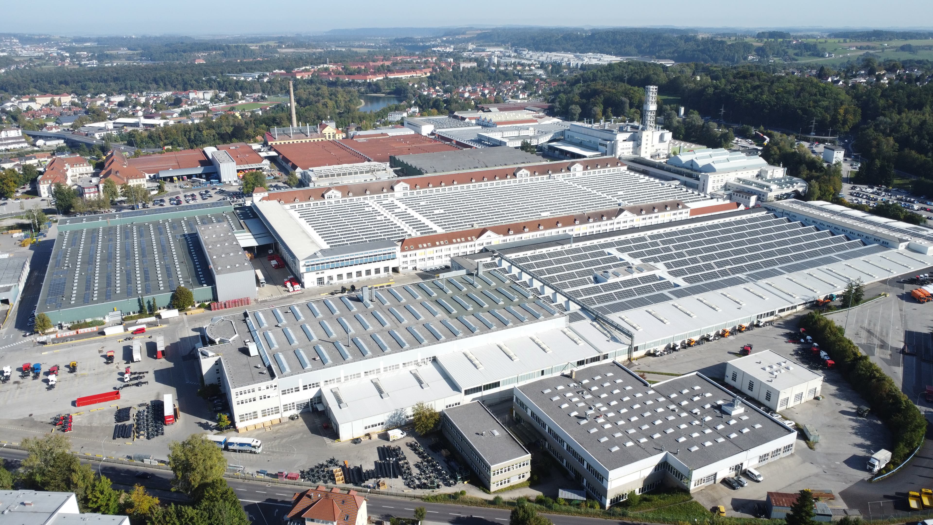 Aerial view of a large industrial complex with multiple buildings, solar panels on rooftops, surrounding greenery, and a neighboring residential area.