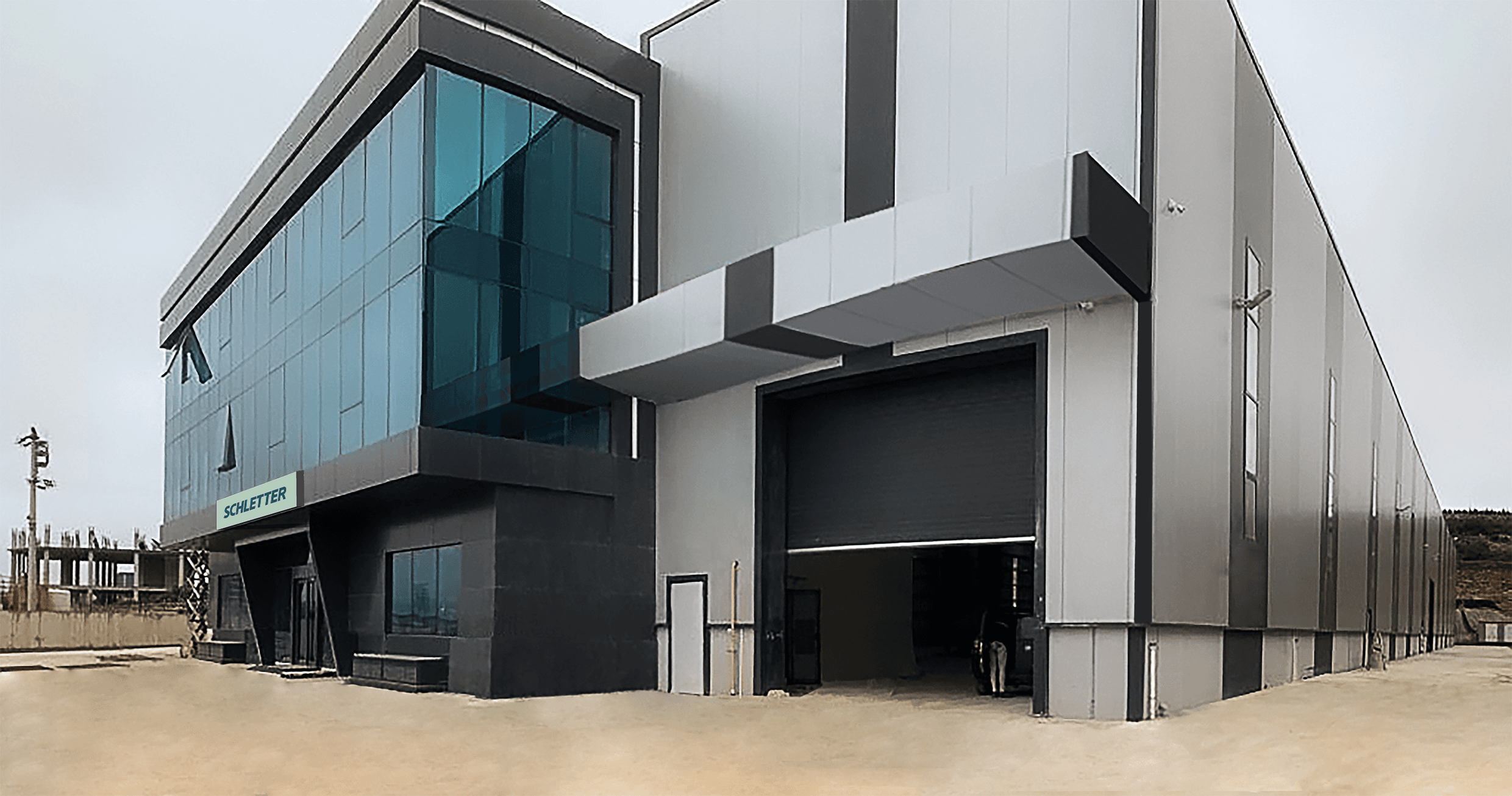 A modern industrial building with a glass-fronted facade and large loading bay. The exterior features a combination of dark and light gray panels. The entrance area has a sign labeled "SAFETY.