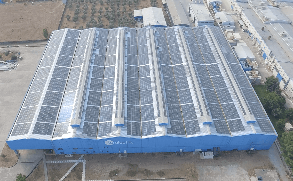 Aerial view of a large industrial building with a blue roof covered in solar panels, labeled "electric." The surrounding area includes other industrial buildings and open land.