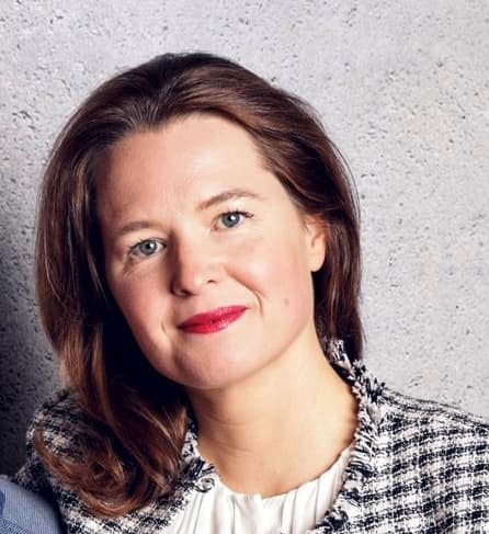 Woman with shoulder-length brown hair, wearing a white blouse and a checkered blazer, posing against a light grey textured background.