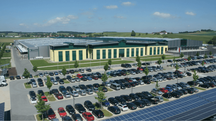 Aerial view of a large industrial building with a solar panel array on the roof, surrounded by a cars-filled parking lot and green landscape.