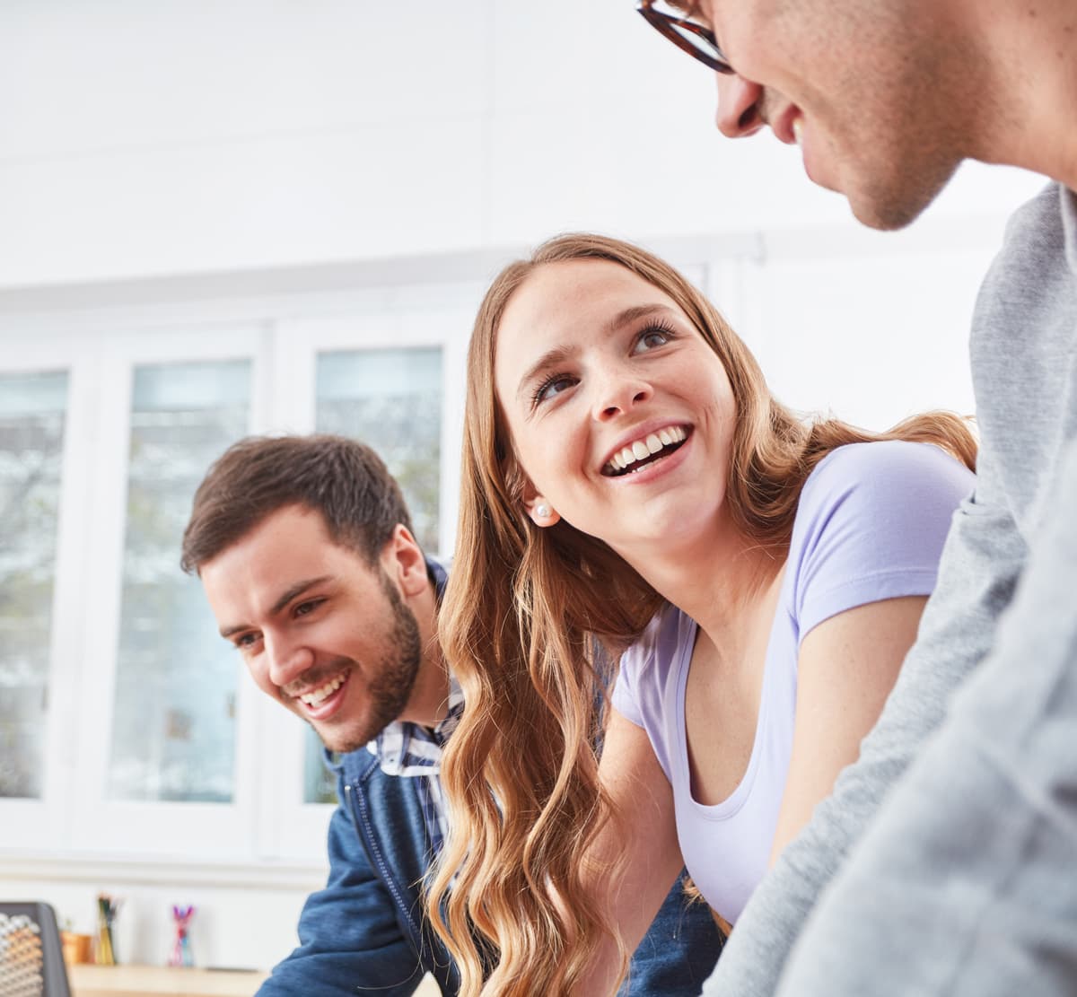 Three people are engaged in a lively conversation, smiling and sitting closely together in a bright room with large windows.