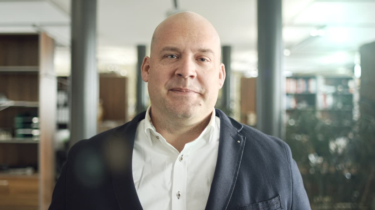 A bald man wearing a white shirt and dark blazer stands in an office with shelves and plants in the background.