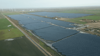 Aerial view of a large solar farm comprising numerous solar panels arranged in rows within a vast, open field. Wind turbines are visible in the distance under a clear sky.