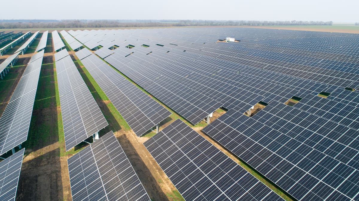 Aerial view of a large solar farm with numerous rows of solar panels installed on a wide expanse of flat land. Trees can be seen at the horizon.