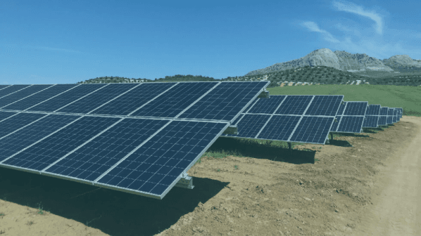 Rows of solar panels installed on a grassy field with mountains in the background under a blue sky.