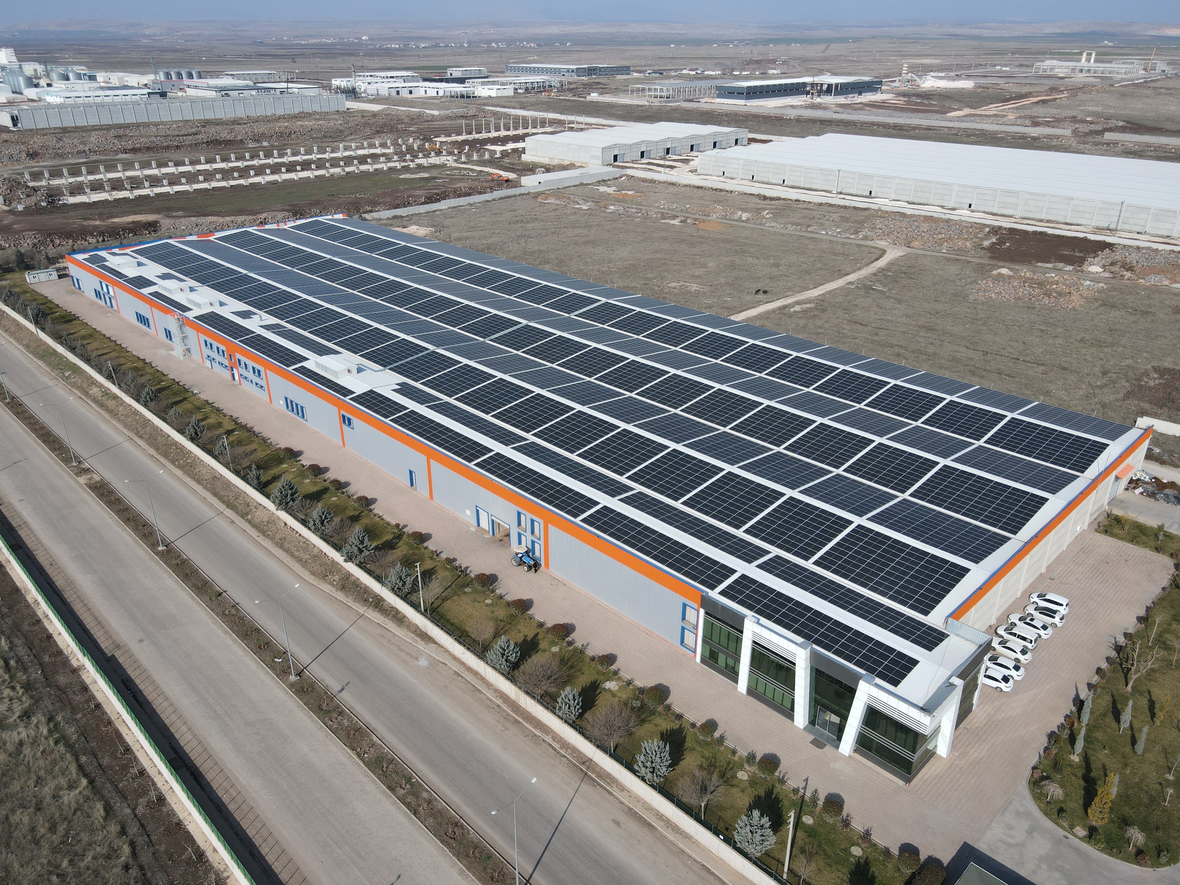 Aerial view of a large industrial building with a solar panel-covered roof, situated in an expansive, sparsely populated area with other industrial structures visible in the background.