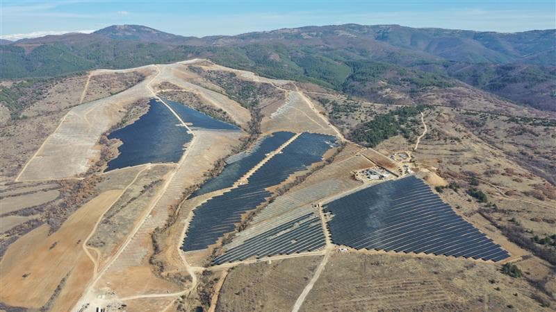 Aerial view of expansive solar panel arrays on a barren hillside surrounded by seemingly forested areas.