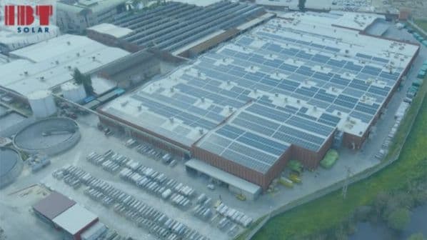 Aerial view of an industrial facility with a large number of solar panels installed on the rooftops. Solar panels and storage are present in the foreground. The image includes the logo "IBF Solar.